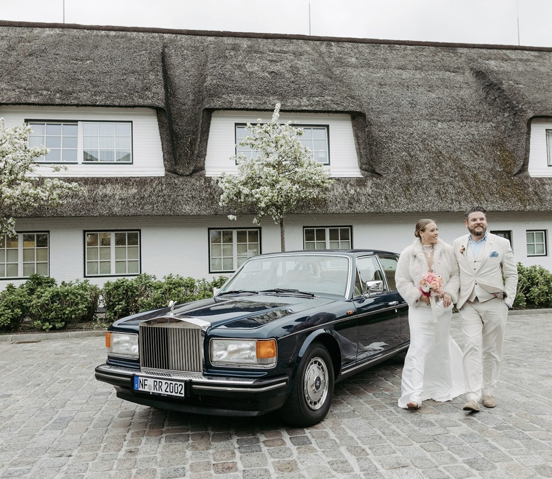 Brautpaar neben einem dunklen Rolls-Royce vor einem reetgedeckten Haus